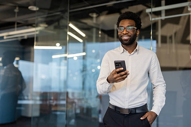 istock Successful businessman with phone in hands standing inside office at workplace. Man smiling contentedly looking out window, financier investor, using app on smartphone 2220277643