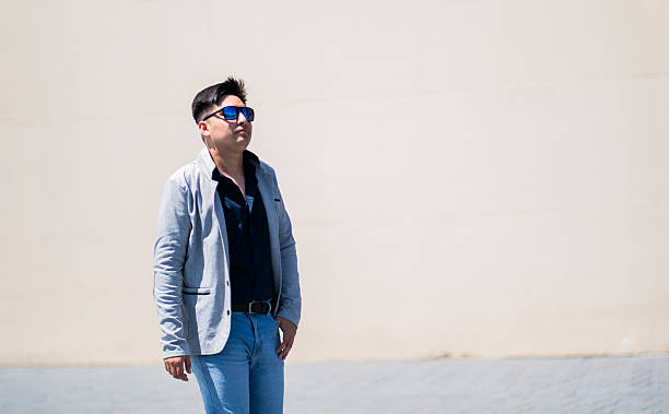 Portrait of a young man wearing light jacket, dark shirt and blue sunglasses, posing confidently in front of a plain, light background. Image representing concepts of leadership, entrepreneurship, executive fashion and contemporary urban style.