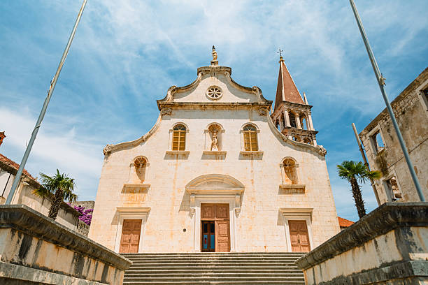 A striking frontal view of a historic baroque-style church on Brac Island, Croatia, featuring symmetrical architecture, carved statues, wooden doors, and a tall bell tower rising into the vibrant summer sky. The image captures the charm of traditional Dalmatian religious heritage, ideal for themes of travel, architecture, spirituality, and European history.