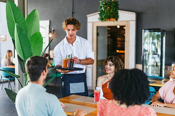 Group of young adults enjoying non-alcoholic beverages in a lively restaurant setting. A waiter serves drinks as friends laugh and connect. The atmosphere is casual and friendly, showcasing togetherness and enjoyment.
