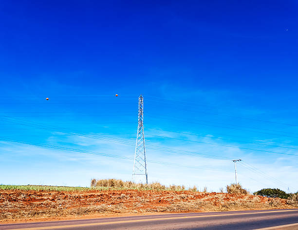 Low-angle view of a high voltage electricity transmission tower under a clear blue sky. The metal structure supports multiple power lines, symbolizing energy distribution and infrastructure. Sunlight highlights the details of the tower, creating a modern industrial feel.