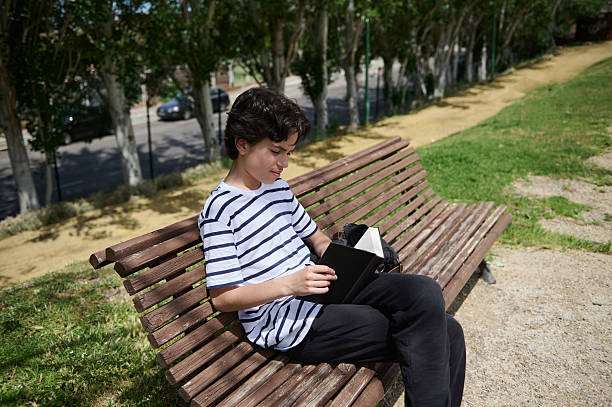 istock Young Boy Peacefully Reading a Book on a Wooden Bench in a Serene Park Setting 2220309291