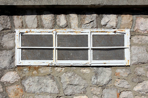 A rectangular window with a rusted metal grille and mesh is set in a rough stone wall, showing signs of age and weathering, with sunlight casting shadows on the textured surface