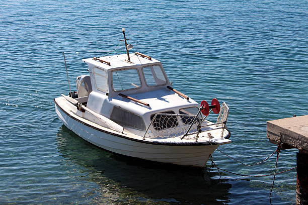 A compact white motorboat with a cabin is tied to a concrete pier, floating on calm blue water with gentle ripples, set against a clear sky, evoking peaceful recreation and maritime travel