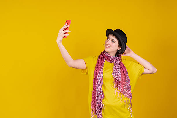 Young woman wearing a hat and scarf takes a selfie with a smartphone, smiling and looking at the screen against a vibrant yellow background