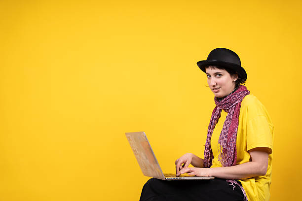 Young woman wearing a bowler hat and scarf using a laptop computer while looking at the camera against a vibrant yellow background