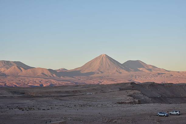 A wide-angle landscape captures the majestic Licancabur Volcano, a prominent stratovolcano on the border of Bolivia and Chile, bathed in the warm, soft light of either sunrise or sunset. The lower slopes of the volcano and the surrounding arid terrain of the Atacama Desert are painted in hues of pink and orange, contrasting with the pale blue sky. The foreground features rocky, desert ground with subtle tire tracks, and a few vehicles are visible, providing a sense of scale to the vast, open landscape. This image beautifully conveys the stark beauty and dramatic lighting of the high-altitude desert. Ideal for travel and tourism campaigns, geological themes, environmental awareness, or any commercial project emphasizing unique global landscapes and adventure