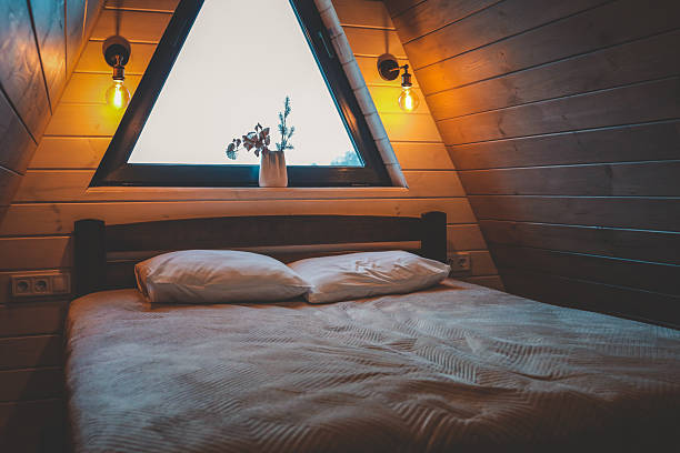 istock Warm bedroom interior in wooden A-frame cabin, bed with pillows, triangular window with forest view, cozy lighting with wall lamps, natural wood decor 2220321687