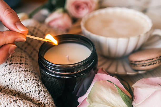 istock A cozy scene with a hand lighting a candle using a matchstick, surrounded by soft beige fabric, a cup of frothy coffee, pink roses, and a macaron. Warm and inviting atmosphere 2220332793