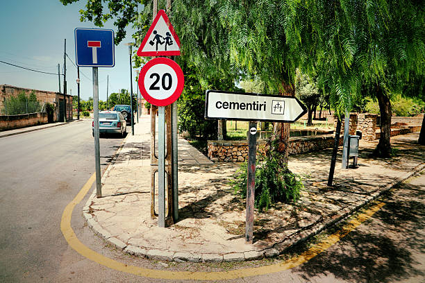 Traffic signs near a sunlit street in Mallorca, Spain, include a dead-end sign, a triangular pedestrian warning, a circular speed limit of 20 km/h, and a directional arrow towards a cemetery (\