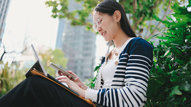 A Gen Z woman focuses on a tablet while sitting in the park, symbolizing flexibility in learning and working.  remote work, online class, digital workspace, tablet use, tech-savvy student, greenery, education tech.