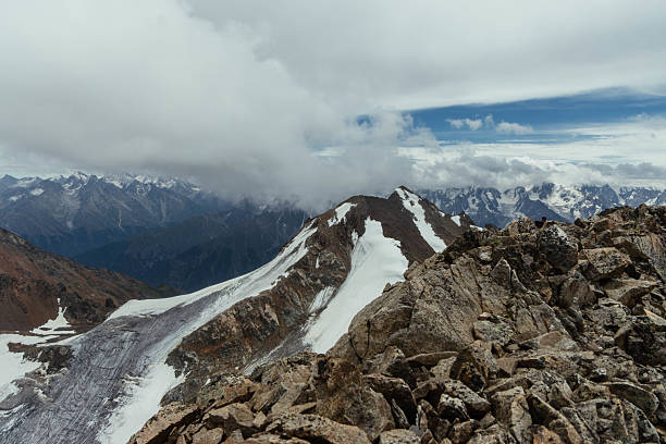 Snow-covered peaks rise above rugged rocks with a backdrop of dramatic clouds in the Andes Mountains. landscape features glacial areas and distant mountain ranges under a blue sky.