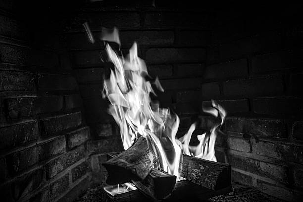 A black and white image captures a fireplace with flames dancing around wooden logs, creating a dramatic contrast against the textured brick background.
