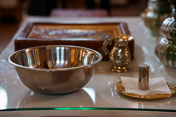 A polished silver bowl sits on a glass table alongside a decorative pitcher, small candle, and scripture, conveying a serene atmosphere for a Christian baptismal rite