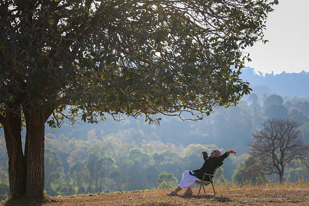 A person is seen relaxing outdoors under a large tree on a dry grassy field, with a mountain and forest landscape in the background. The individual is leaning back in a chair with an arm raised, conveying joy and freedom amidst nature.