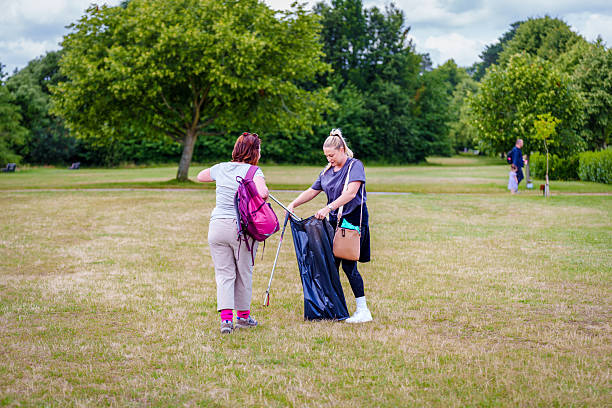 This image captures a group of people actively engaged in an outdoor activity,  a community volunteer project or environmental work. They are raking  dry grass in a sunlit, grassy area surrounded by lush green trees. The individuals are dressed in casual, comfortable clothing suitable for outdoor work, and they appear to be enjoying the task. The bright, natural setting and the teamwork evident in their efforts create a positive and productive atmosphere. This scene suggests a communal effort towards maintaining or improving a natural space, fostering a sense of environmental stewardship and collaboration.