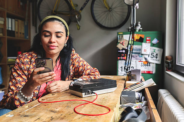 young happy woman connecting modern smartphone with retro tape recorder at wooden table in workshop