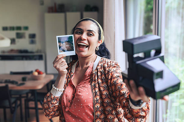 young smiling woman taking selfie with retro instant camera in living room