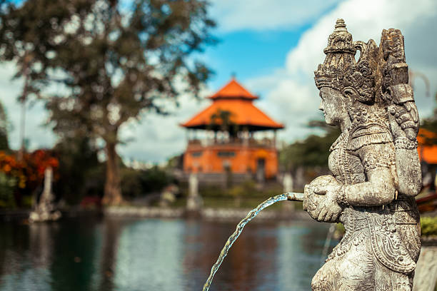 Close-up of ornate stone fountain statue in Tirta Gangga, Bali, with traditional orange-roofed temple and reflective pond in background.