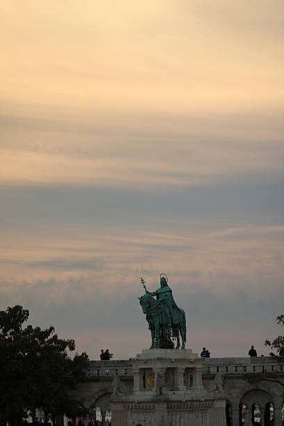 A distant shot of the statue of King Stephen I of Hungary set against a vivid blue sky scattered with smooth white and orange clouds. The serene backdrop highlights the monument’s historic significance and majestic presence.