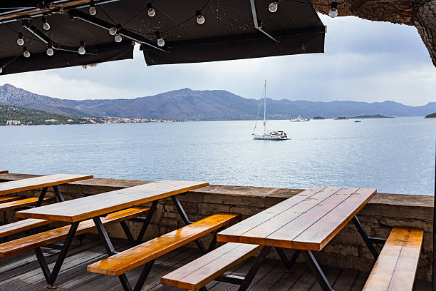 Charming seaside dining area in Korčula, Croatia, featuring wooden tables and benches overlooking the Adriatic Sea. A sailboat floats peacefully on the calm water, framed by potted plants and distant island hills under a cloudy summer sky. The warm textures of the wood and stone contrast with the refreshing maritime backdrop, creating a serene Mediterranean lifestyle and travel scene, ideal for hospitality, leisure, and coastal tourism concepts.