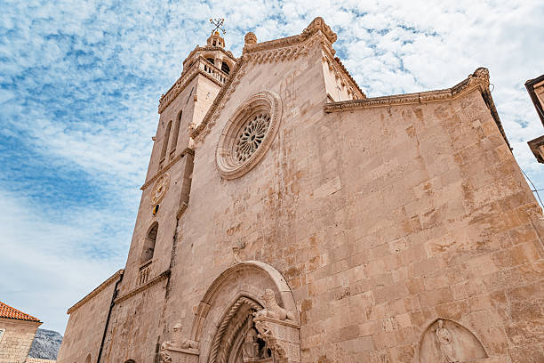 Low-angle view of St. Mark’s Cathedral in Korčula, Croatia, showcasing the impressive bell tower, detailed clock, and rose window set against a bright summer sky. The architecture represents a blend of Romanesque and Gothic influences common in Dalmatian island towns. This striking image is ideal for editorial use, travel promotions, cultural heritage campaigns, and Mediterranean tourism content.