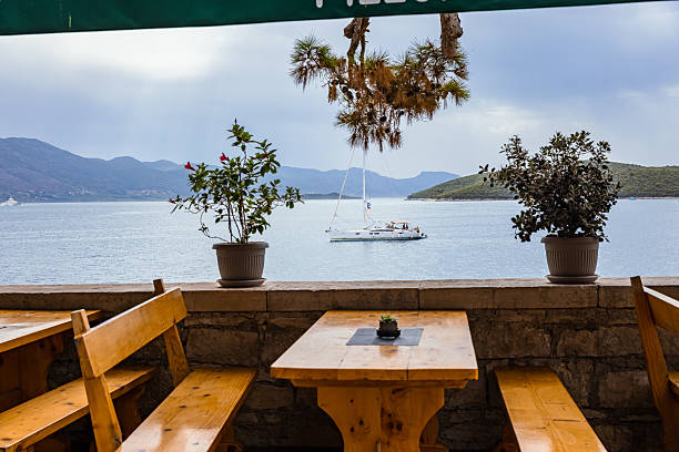 Charming seaside dining area in Korčula, Croatia, featuring wooden tables and benches overlooking the Adriatic Sea. A sailboat floats peacefully on the calm water, framed by potted plants and distant island hills under a cloudy summer sky. The warm textures of the wood and stone contrast with the refreshing maritime backdrop, creating a serene Mediterranean lifestyle and travel scene, ideal for hospitality, leisure, and coastal tourism concepts.