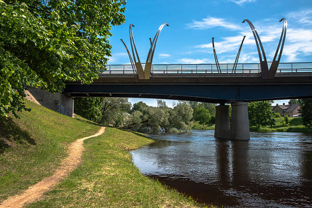 istock Modern pedestrian bridge with artistic metal arches spanning over a calm river, viewed from a grassy riverside path under a blue summer sky 2220395816