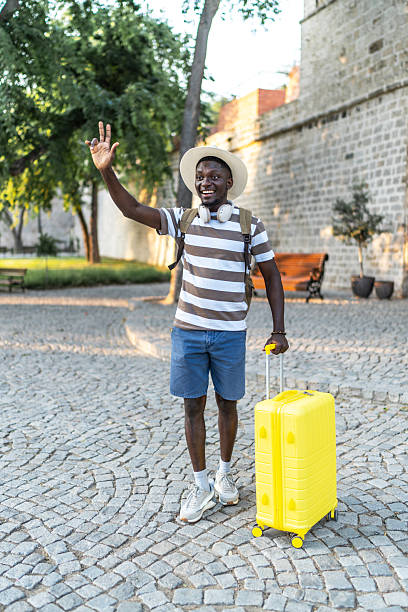 istock Young black tourist waving while carrying a yellow suitcase 2220404720