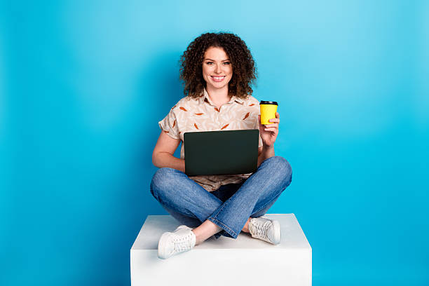 istock Happy young woman with curly hair using laptop and drinking coffee while sitting on a platform with a bright blue background 2220407859