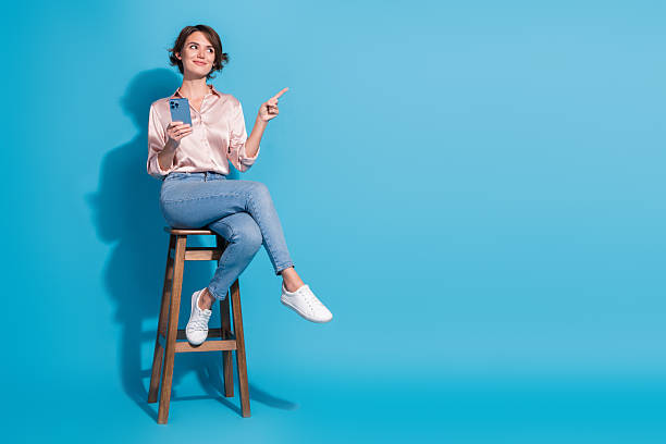 istock Young woman in elegant satin blouse joyfully pointing finger while holding smartphone against a vibrant blue background 2220411497