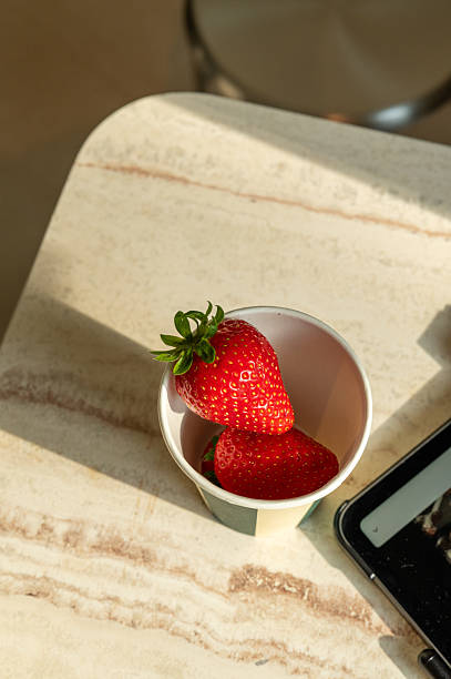 Two ripe strawberries sit in a disposable cup on a marble table, illuminated by natural light