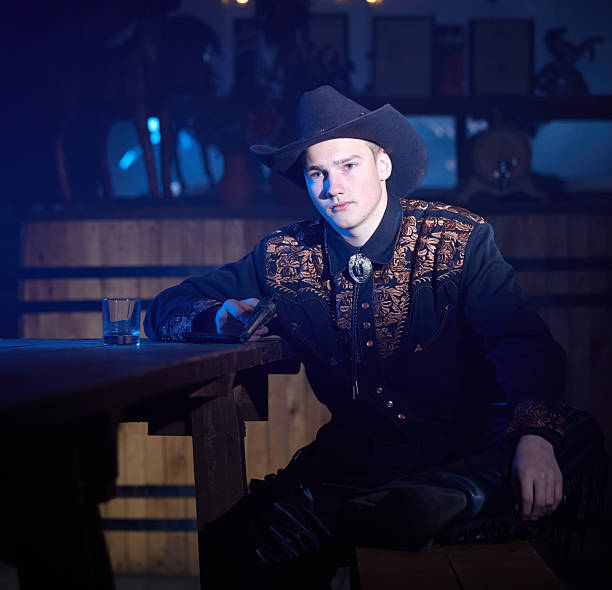 Young handsome cowboy is sitting at table in saloon, looking at the camera