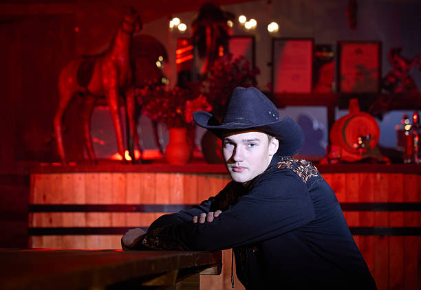 Young handsome cowboy is sitting at table in saloon, looking at the camera