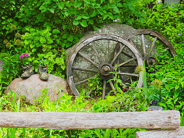 Charming rustic garden decoration featuring old wooden wagon wheels, vintage boots, stones and vibrant green plants. Perfect for gardening, countryside living, and rustic design projects.