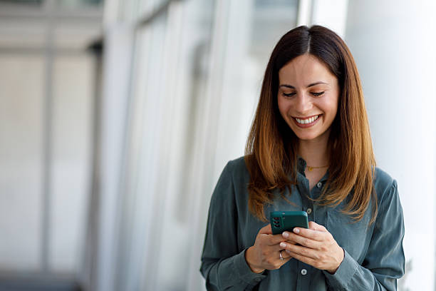 istock Happy businesswoman smiling while using a mobile phone, standing confidently in a modern office space filled with natural light 2220423597
