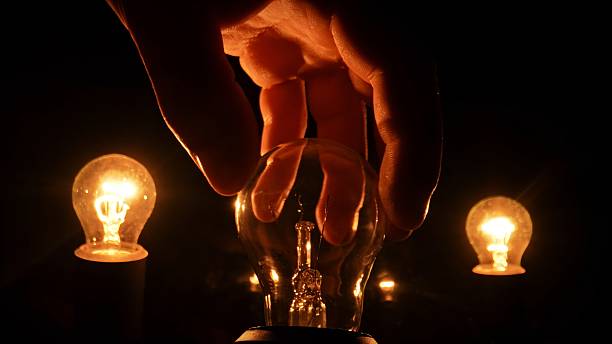 A hand gently touches an unlit light bulb surrounded by glowing bulbs in a dark environment. The image conveys contrast, creativity, and the concept of potential or inspiration.