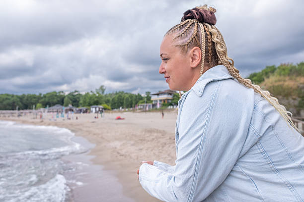 A woman in a black and light denim jacket leans against the railing of a bridge. The water is calm with no waves, and the sky is gloomy. Off season!