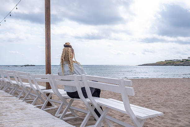 A woman in a black and light denim jacket sits on a white bench on a deserted beach, her back to the camera, looking out to sea. The water is calm with no waves, and the sky is gloomy. Off season!