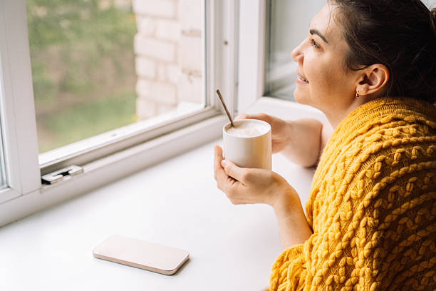 istock Woman smiling while holding coffee and looking out the window, enjoying tech-free time 2220444940