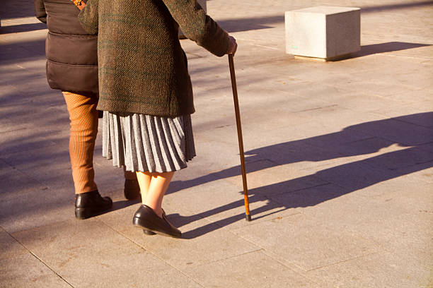 Two women walking on pedestrian zone, senior one with walking cane. Sunny weather in wintertime, Alcalá de Henares, Madrid,  Spain.