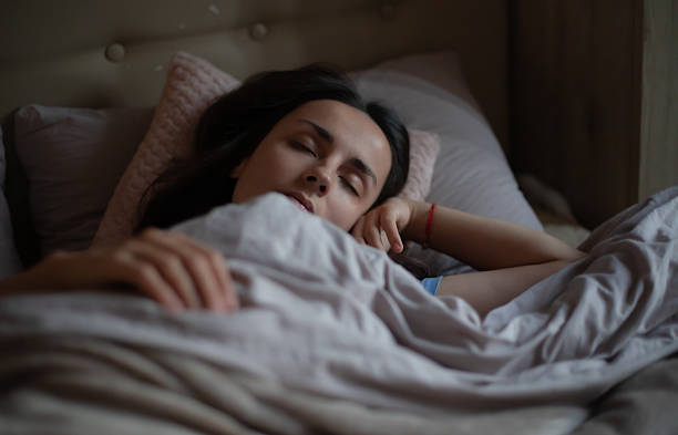 istock Young woman peacefully sleeping in bed under a cozy blanket, resting her head on a soft pink pillow in a dimly lit bedroom with a calm, serene atmosphere. Ideal for sleep or wellness themes. 2220451559