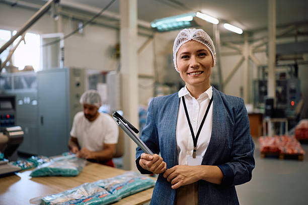 Female food factory worker holding a tray of freshly baked cookies, smiling in an industrial bakery setting