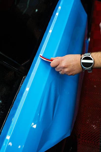 istock Vehicle wrap detailing involves squeegee strokes for seamless, wrinkle-free adhesion. Detailing expert uses red squeegee wrapper to flatten blue PPF wrap on car surface. 2220466748