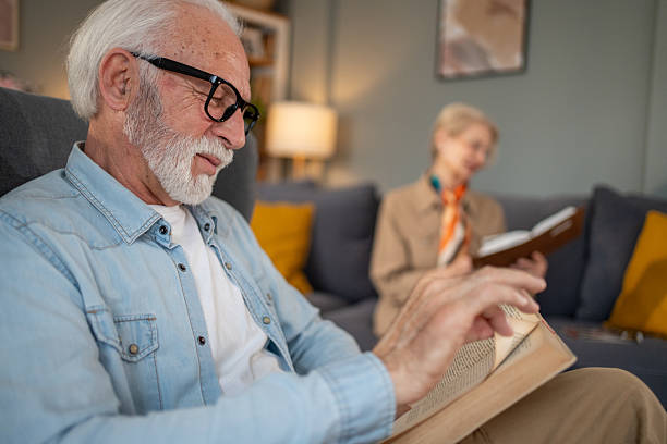 Senior man reading a book while sitting on sofa at home with his wife