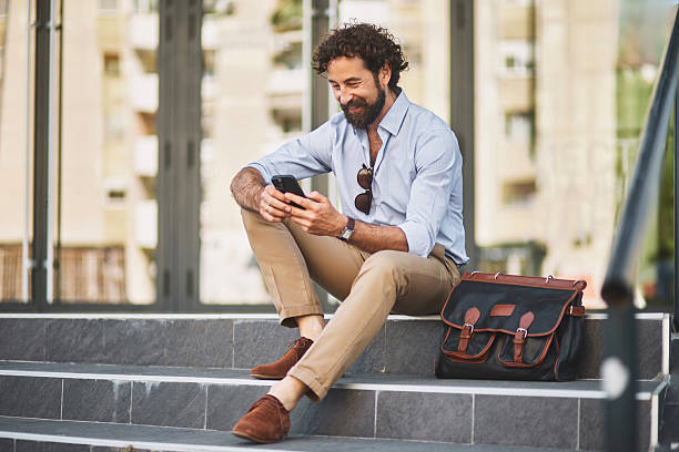 istock Businessman sitting on stairs using smartphone and smiling 2220469695