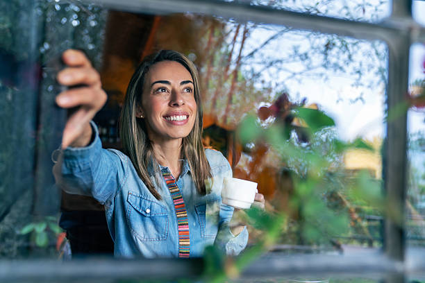 istock Happy woman looking through the window while drinking coffee in a log cabin 2220472599