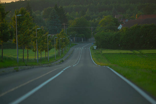 A car is steadily driving down a winding country road that is beautifully surrounded by lush trees and vibrant green grass on both sides