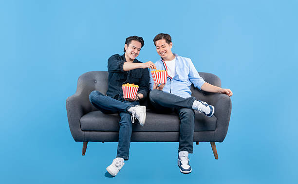 istock Friends sharing popcorn on sofa against a vibrant blue backdrop, smiling and enjoying quality time together in a relaxed and casual setting. 2220494971