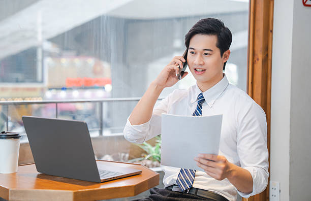 istock Professional working in a cafe talking on his phone reviewing a document and using a laptop 2220502055